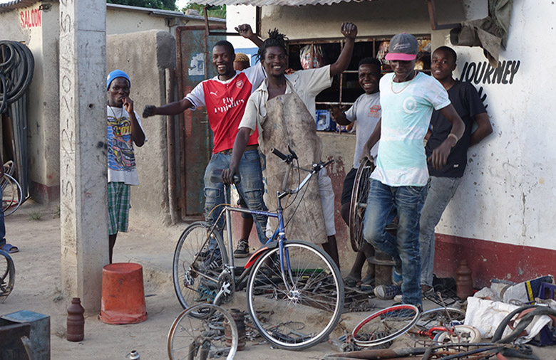 Devant un bâtiment et derrière un vélo se tiennent quelques jeunes hommes de très bonne humeur.
